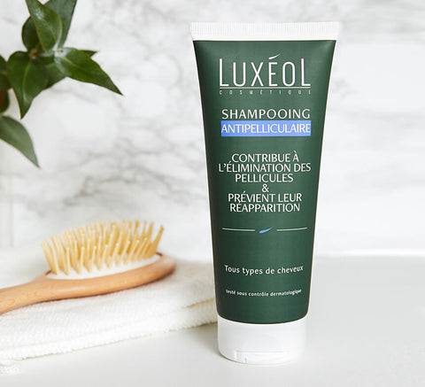Tube of dark green Luxéol anti-dandruff shampoo with white cap on bathroom counter next to hairbrush and white towel.